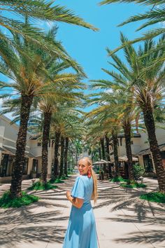 a woman in a blue dress is standing under palm trees and looking up at the sky