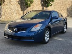 a blue nissan car parked in front of a wall with trees on it's side