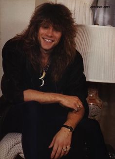 a man with long hair sitting in front of a lamp and smiling at the camera