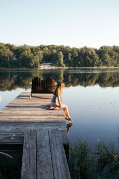 a woman sitting on a dock next to a body of water with trees in the background