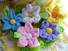 four different colored flowers in grass on a cloth covered surface with yellow, blue, pink and green fabric