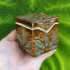 a hand holding a small decorative box in front of a green leafy background,