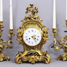 an ornate clock with candles in front of it on a gray background and two white candles behind it