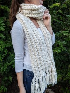 a woman wearing a white knitted scarf with tassels on the ends, standing in front of some bushes