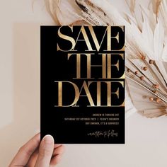 a hand holding up a black and gold save the date card with feathers on it