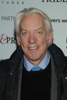 an older man in a black jacket and blue tie smiles at the camera while standing next to a white wall