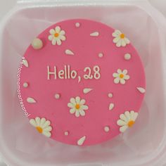 a pink cake with white daisies and the words hello, 28 on it is in a plastic container