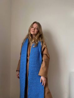 a woman standing in front of a white wall wearing a blue knitted coat and brown sweater