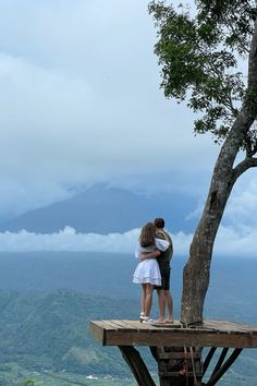 two people standing on top of a wooden platform next to a tree with mountains in the background