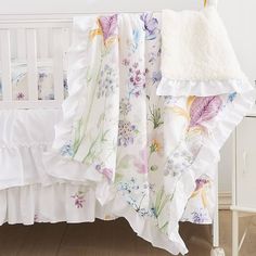 a white crib with floral bedding and blankets on it's sides, next to a dresser