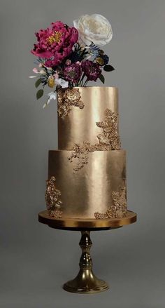 a three tiered cake with gold icing and flowers on the top is shown in front of a gray background