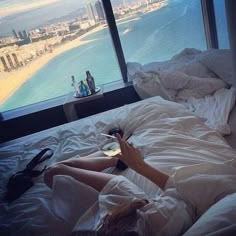 a woman laying in bed looking out the window at an ocean and cityscape