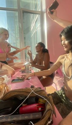 three women in bathing suits sitting at a table with food and drinks, one holding a cell phone