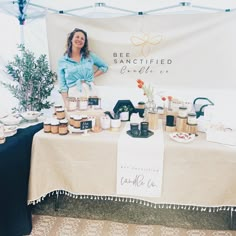 a woman standing behind a table with jars and cups on it, in front of a sign that says bee sanctified edible co