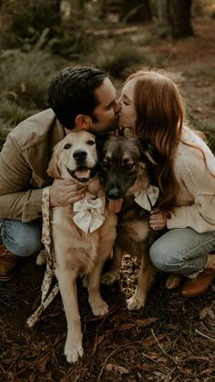 a man and woman kissing while holding two dogs in the woods with their faces close to each other