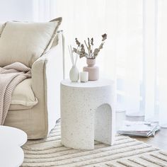 a white table with vases and flowers on it in front of a chair next to a window