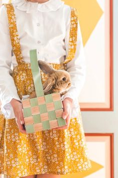 a woman holding a basket with a rabbit in it's lap while wearing a white shirt and yellow dress