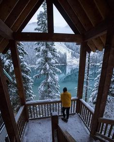 a person standing at the top of a wooden porch looking out over a lake and snow covered trees