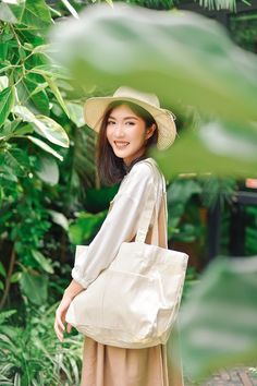 a woman in a hat and dress carrying a large white tote bag while standing next to green plants