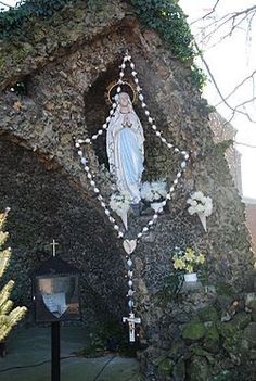 a statue of the virgin mary in front of a stone wall with flowers on it