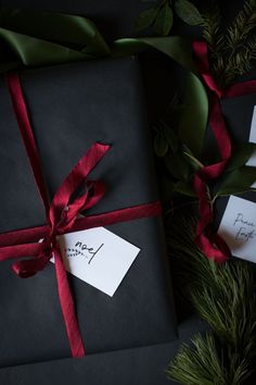 two wrapped presents with red ribbon and tags on them next to some evergreen branches,