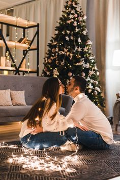 a man and woman sitting on the floor in front of a christmas tree with lights