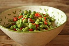 a white bowl filled with green and red vegetables on top of a wooden table next to a spoon