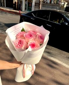 a woman holding a bouquet of pink roses on the side of the road in front of a black car