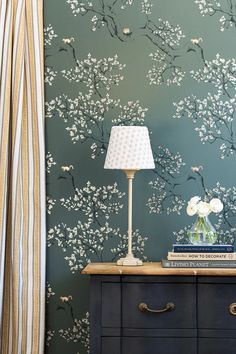 a nightstand with a lamp and flowers on it in front of a wallpapered background