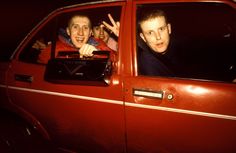 two men sitting in the back of a red car with their hands up and smiling