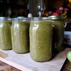 four jars filled with green smoothie sitting on top of a wooden cutting board next to an apple