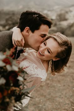 a bride and groom embracing each other in front of a mountain range with flowers on the ground