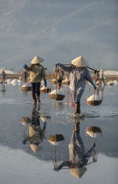 two people walking on the beach carrying sand in their hands and wearing straw hats, with mountains in the background