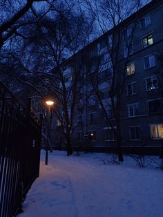 a street light in the middle of a snow covered park at night with apartment buildings behind it