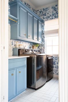 a kitchen with blue cabinets and white tile flooring is seen from the doorway to the washer and dryer