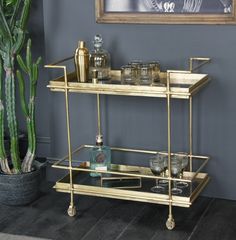 a gold bar cart with glasses and bottles on it next to a potted cactus