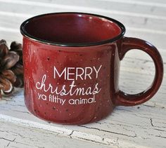 a red coffee mug with merry christmas ya filthy animal written on it next to some pine cones