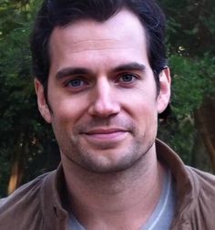 a close up of a person wearing a brown jacket and smiling at the camera with trees in the background