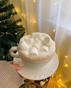 someone holding a cup of hot chocolate with marshmallows on top in front of a christmas tree