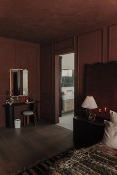 a bedroom with red walls and wooden flooring has a bed, desk, mirror and lamp