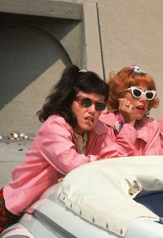 two women sitting on the back of a car talking on their cell phones while wearing sunglasses