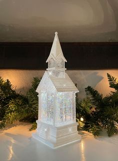 a lit up white church on top of a table next to evergreens and lights