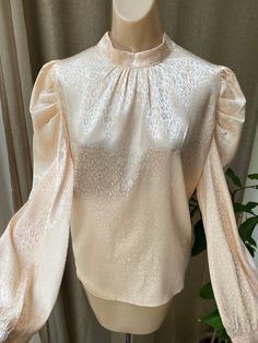 "This is a wonderful blouse for the lady who can't pass up 'just one more' great blouse. Size tag is Large but I think it can work for a lady who wears a size M as well as the style isn't meant to be tight on the body. Fabric is 97% polyester and 3% spandex, a delicious combination that feels and drapes beautifully. Never worn and perfect. Even an extra fabric covered button in a side seam. Color is hard to describe - a soft slightly antiqued yellow with a slight sheen from the embossed pattern. Cream Blouse With Blouson Bishop Sleeves, Cream Blouse With Puff Blouson Sleeves, Cream Blouse With Blouson Puff Sleeves, Elegant Beige Blouse With Blouson Sleeves, Feminine Party Blouse With Bishop Sleeves, Elegant Cream Tops With Gathered Sleeves, Elegant Cream Top With Gathered Sleeves, Feminine Evening Blouse With Lantern Sleeves, Feminine Bishop Sleeve Party Blouse