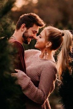 a man and woman standing next to each other in front of pine trees at sunset