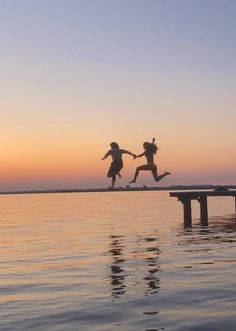 two children jumping off a dock into the water at sunset or dawn with their arms in the air