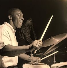 a black and white photo of a man playing drums with another man in the background