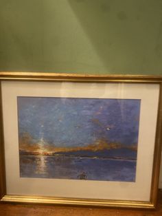 a painting hanging on the wall next to a wooden table with a white and gold frame