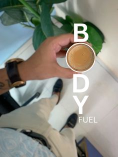 a person holding a cup of coffee with the words body fuel on it and a plant in the background