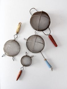 three metal pots with handles and wire hanging from the sides on a white wall next to two different colored kitchen utensils