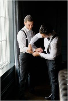 two men standing next to each other in front of a window wearing suspenders and ties